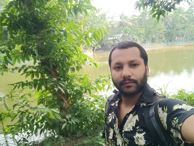 A man with a beard takes a photo, holding his backpack while standing near a tree. The background shows a lake surrounded by trees, creating a serene setting for the photograph. In the caption provided, there are mentions of apps, like Nomisma Network Bullish or @perx_trade, and the theme is related to sleep.