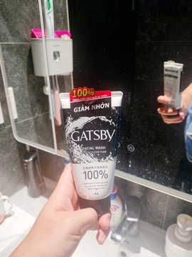 A hand holding a black and white tube of "GATSBY GIAM NHON" facial wash marked "100%" in a bathroom setting with a mirror reflection.