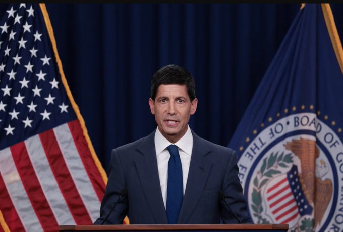 A man in a dark suit and blue tie stands at a podium delivering remarks, with an American flag on the left and the Federal Reserve Board of Governors seal on the right.