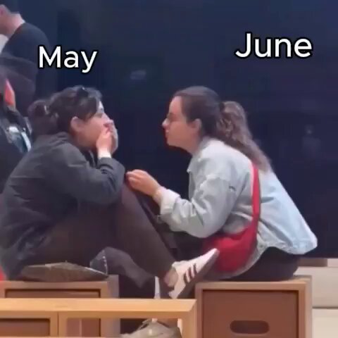 The image shows two women sitting next to each other with their legs crossed, possibly having a conversation or enjoying some time together. They are positioned near a wooden table that has a red bag placed on top of it. The background includes a TV screen displaying the text "May is shaping up to be a very exciting month."
