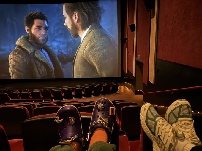 A view from inside a movie theater showing someone's feet propped up in front of a large screen displaying two men in conversation, with a pair of chunky sneakers and purple shoes visible.