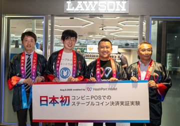 Four men wearing happi coats and medals stand inside a Lawson store holding a banner that reads 'JAPAN-POSでの ステーブルコイン決済実証実験' (Japan's first stablecoin payment experiment at Lawson POS).