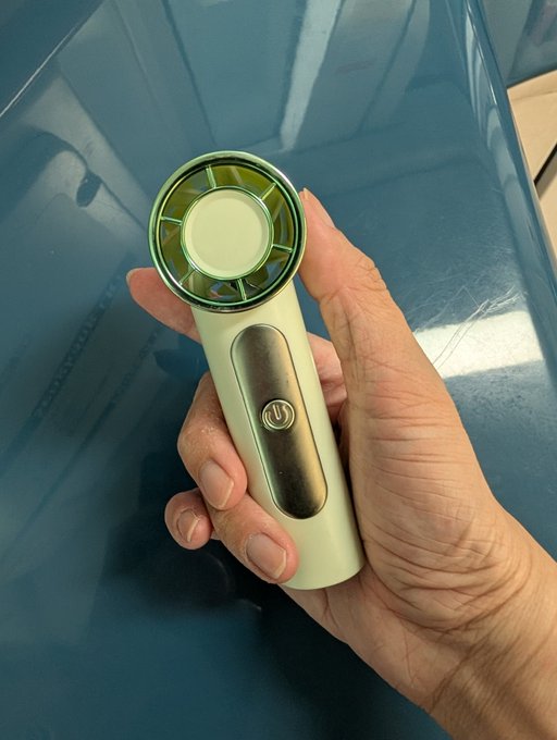 A hand holds a mint green handheld electric fan with a silver power button, set against the glossy blue surface of what appears to be a car. The fan serves as essential relief gear for surviving Singapore's notoriously hot and humid weather, especially during crowded events like Token2049 and the F1 race.