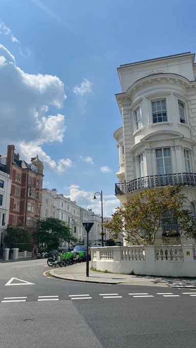 A street view of elegant white and brick Victorian-style buildings on a curved road under a blue sky with scattered clouds.