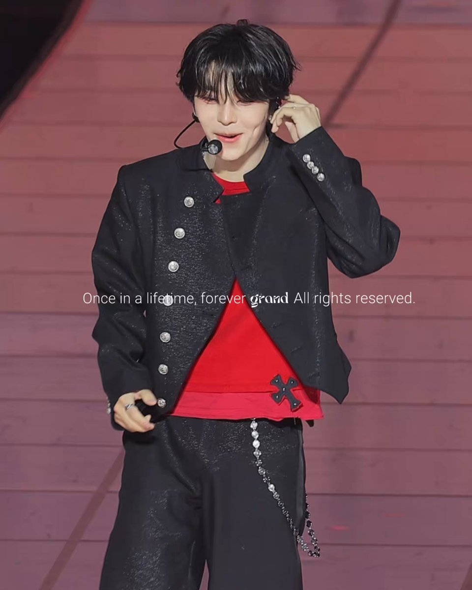 The image features a young man wearing black clothing, including pants and a jacket with buttons down the front. He has short hair and is holding a microphone close to his ear while standing on stage. The background of the photo shows a wooden floor, which complements the overall aesthetic of the scene. There are also some words written in the image that read "260614 롱마트만세 #XINLONG #鑫隆 #씬롱 #알디원".