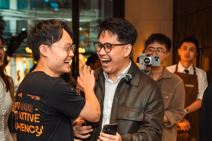 The image shows a group of people gathered together, with two men standing out as they laugh and share a moment. One man has his arm around another man's shoulder, while both are smiling at each other. The rest of the group appears to be engaged in conversation or enjoying the event. In the background, there is a camera capturing this memorable moment.

The post features an image of Bitcoin Pizza Day with a man holding a cell phone and a pizza. There are also several people present in the scene, including one person wearing glasses who seems to be laughing along with the others. The overall atmosphere appears to be light-hearted and joyful as everyone comes together for this special occasion.