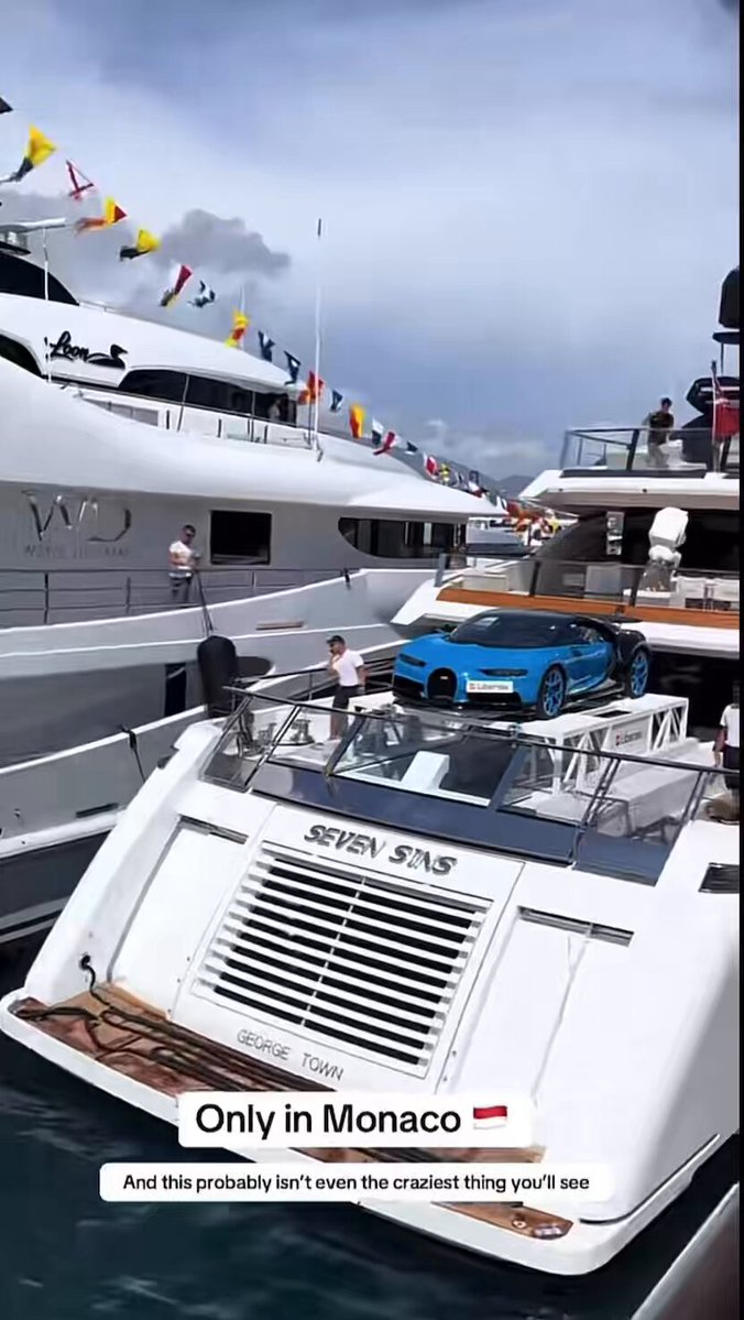 The image shows a large white boat with a blue car on top of it. The boat appears to be docked at a marina or harbor, possibly in Monaco. There are several people around the boat, some standing near the railing while others are closer to the water's edge. A person is also seen walking along the dock area. In the background, there are flags flying from various locations, adding to the festive atmosphere of the scene. The image includes text that reads "Only in Monaco" and "And this product was worth it for you".
