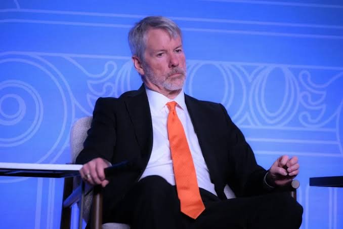 A gray-haired, bearded man wearing a black suit and orange tie sits in a chair against a blue backdrop, appearing to be at a panel discussion or conference.