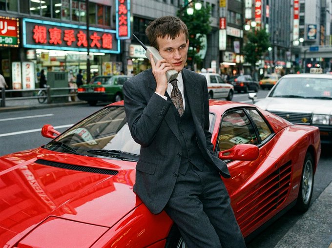A man in a dark suit leans against a red Ferrari sports car while talking on a mobile phone, set against a busy city street with neon signs featuring Japanese text.