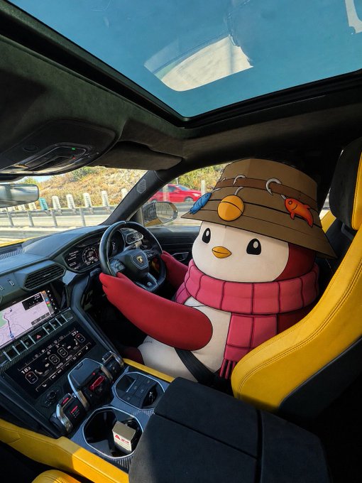 A cartoon penguin mascot wearing a fishing hat and red scarf sits in the driver's seat of a yellow sports car, ready to hit the road. The image, paired with Vietnamese text about an imminent major event for memecoins ("Một điều gì đó lớn lao sắp đến với memecoins"), humorously suggests the mascot is gearing up to drive the next big memecoin rally.