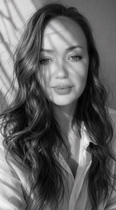 A black-and-white portrait of a woman with long wavy hair, looking directly at the camera, with shadows from a window playing across her face.