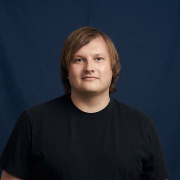 A portrait photo of a young man with light brown hair wearing a black t-shirt, photographed against a dark navy blue studio backdrop.