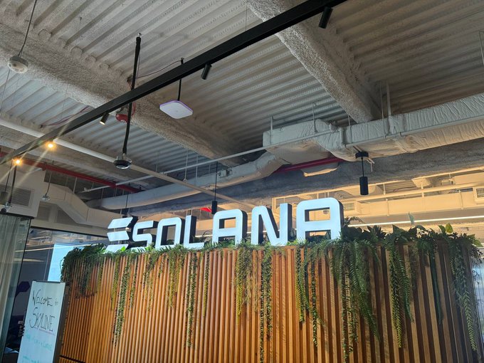 A photograph of a Solana-branded reception area or office lobby featuring the illuminated Solana logo mounted on a wooden slat wall with trailing green plants.