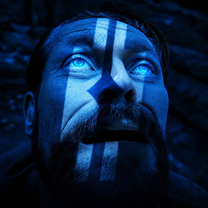A close-up portrait of a bearded man looking upward with glowing blue eyes, his face illuminated by blue light casting vertical stripe shadows across his features.