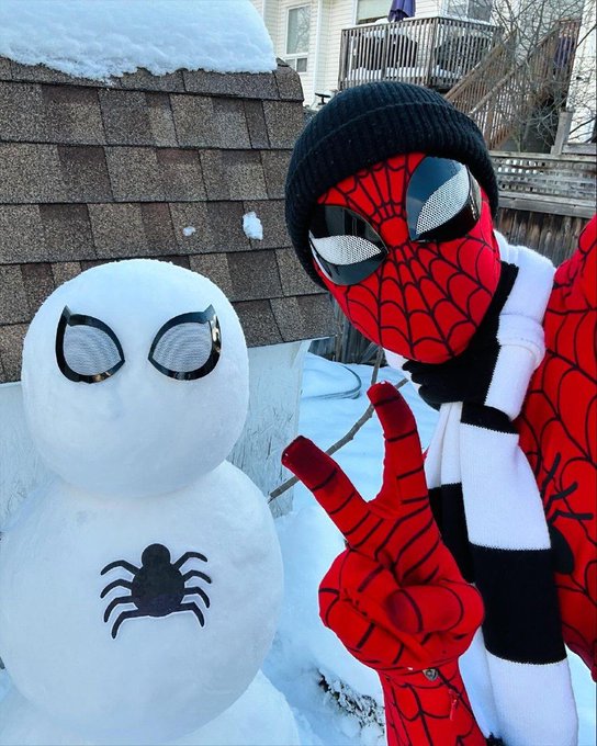 A winter scene featuring a person in a Spider-Man costume and black beanie posing with a peace sign next to a snowman decorated with a spider emblem and eyes.
