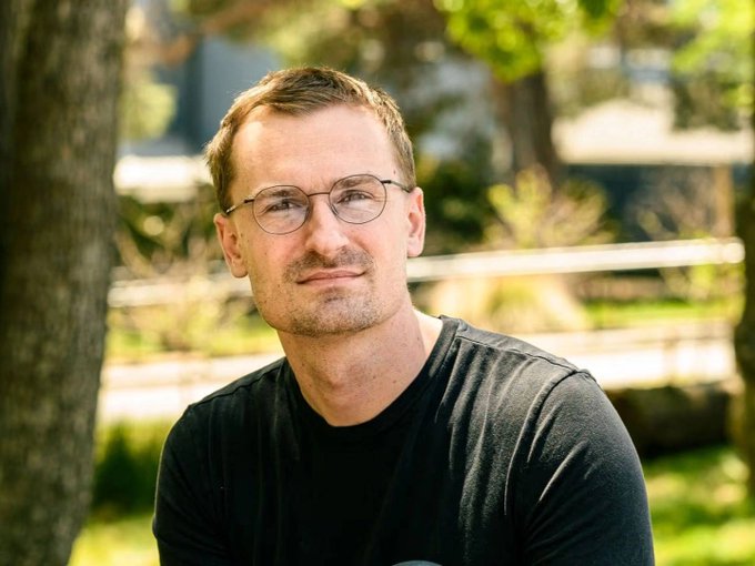 A man with glasses and short brown hair, wearing a black t-shirt, is pictured outdoors in a sunlit, green park-like setting. The portrait pairs with the post's theme of tokenization, visually anchoring the concept of converting real-world identity into digital assets.