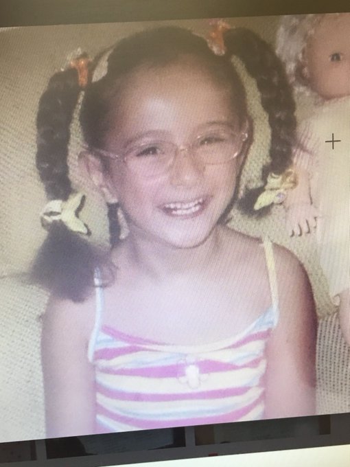 A photograph of a young smiling girl with pigtails and glasses wearing a striped tank top, sitting next to a doll.