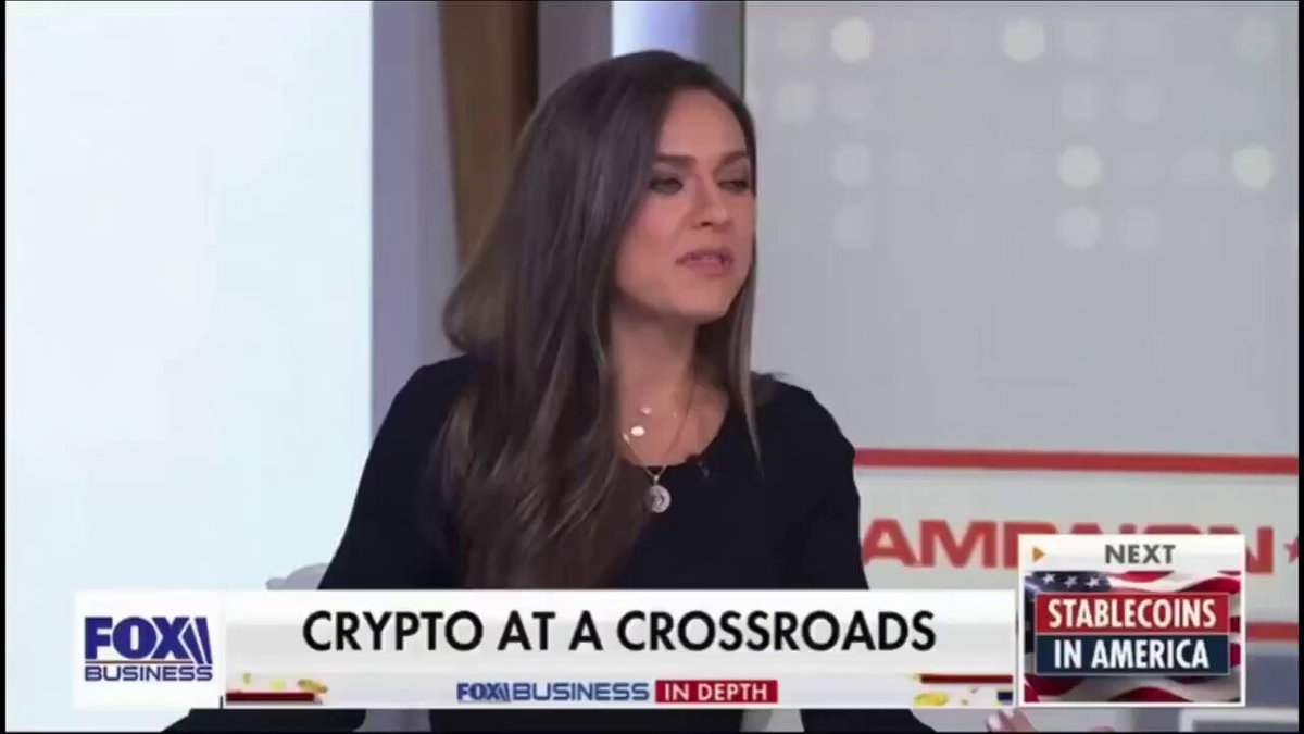 The image shows a woman sitting at a desk, wearing a black top with her hair pulled back into a ponytail. She appears to be talking about Bitcoin on a Fox News show or a similar platform. The background of the image features a white wall with red and blue text that reads "Fiat broke the American Dream. Bitcoin restored it. The world needs Bitcoin. Bitcoin doesn't need the world." This suggests that the woman is discussing how Bitcoin has the potential to change the way people perceive money, as it was created to counter the power of fiat currency like the US dollar.