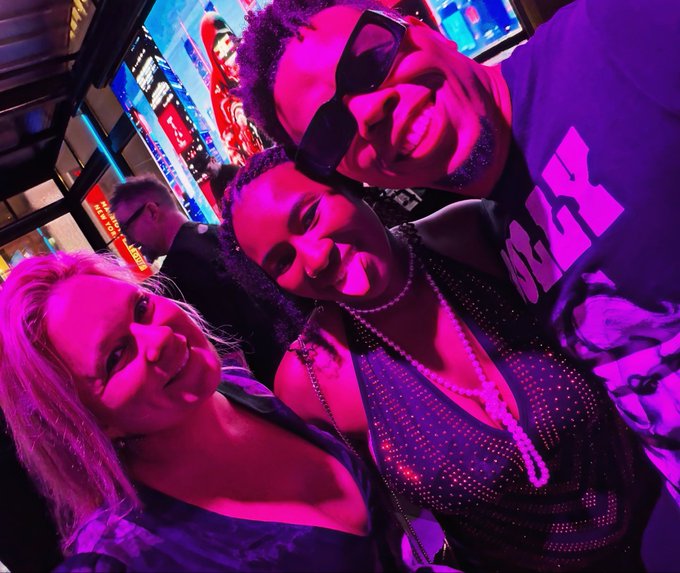 Three friends taking a selfie in a vibrant, neon-lit urban setting with pink and purple lighting, one wearing sunglasses and another in a sequined dress.