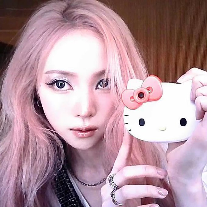 A person with long pink hair poses for a selfie while holding a white Hello Kitty-themed camera case with a pink bow, fitting the caption's description of Narin's first Instagram profile picture. The close-up shot captures her soft makeup, nose ring, and silver rings as she gazes directly at the camera.