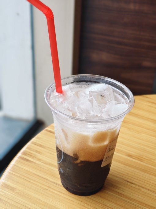 A clear plastic cup filled with iced coffee, cream, and ice sits on a wooden table with a red straw.