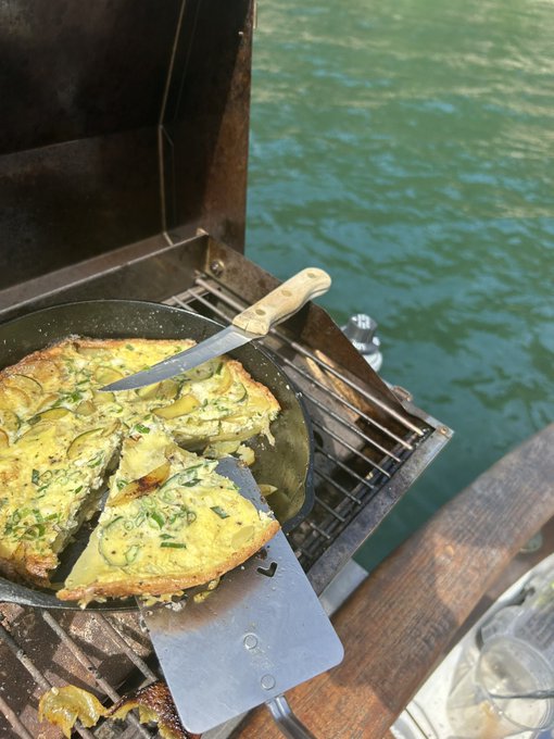 A freshly cooked Spanish tortilla or omelet with potatoes and herbs sits in a cast iron pan on a grill by the sea, with a knife and spatula resting in it.