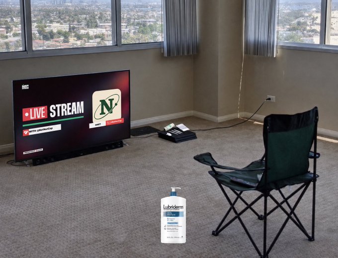 A sparsely furnished room with a TV displaying a 'LIVE STREAM' for a Nebraska football game (N logo), a folding chair, and a bottle of Lubriderm lotion on the carpet.
