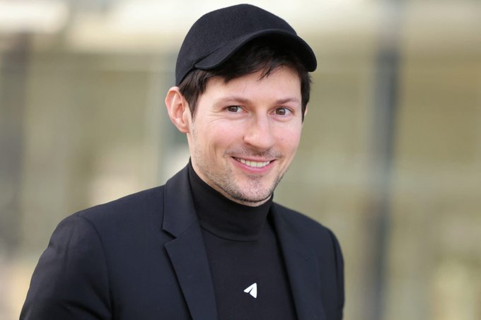 The image features a man wearing a black hat, a black suit jacket, and a black shirt with white text that reads "TON'S NATIVE token will be renamed to Gram". The man appears to be smiling for the camera while posing outdoors in front of a building.