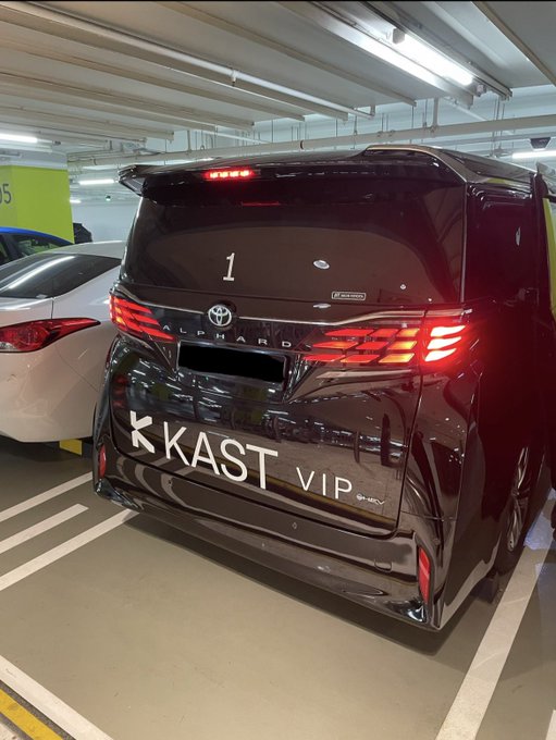 The rear view of a black Toyota Alphard VIP van parked in a parking garage, featuring 'K KAST VIP' lettering across the tailgate and an Alphard badge, with a white car parked next to it.