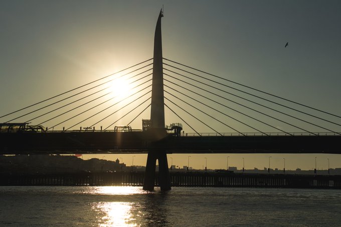A silhouetted cable-stayed bridge is captured at sunset, with the sun shining brightly behind its tall central pylon and reflecting on the water below.