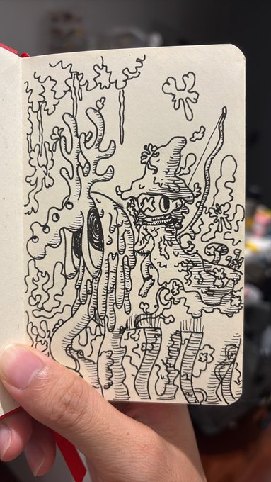 A hand holds open a sketchbook displaying a detailed pen-and-ink drawing of a fantastical landscape featuring coral-like structures, abstract plants, and a central creature-like figure.