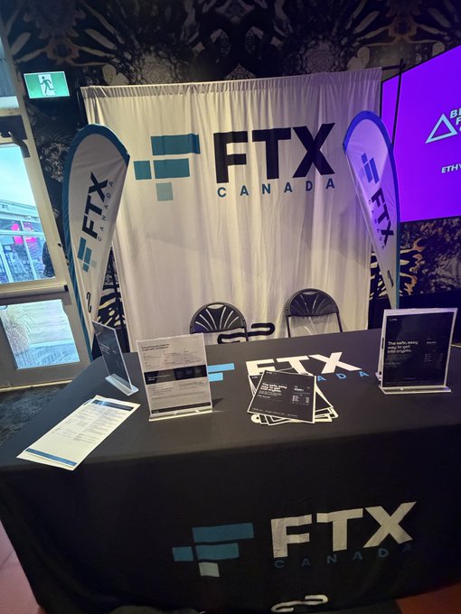 A promotional booth for FTX Canada featuring branded backdrop banners, teardrop flags, and a tablecloth with the FTX Canada logo. Informational pamphlets and cards are displayed on the table, with an exit sign and partial view of another digital display visible in the background.