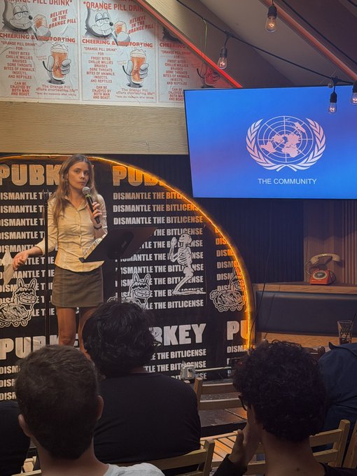 A photograph of a young woman speaking into a microphone at a podium during an event at 'Pubkey', with a screen behind her displaying a UN-like emblem and the text 'THE COMMUNITY'.