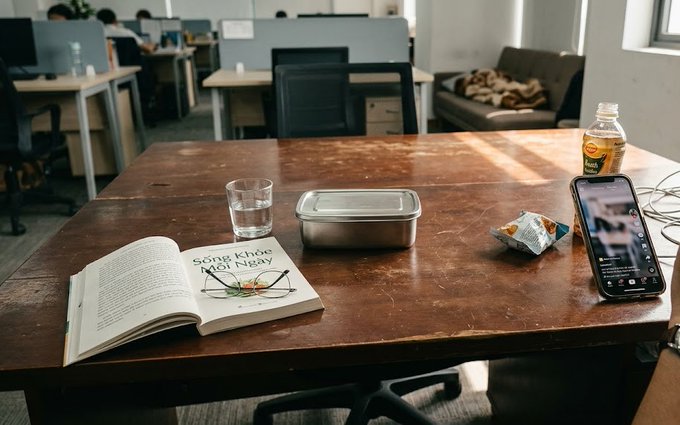 A wooden office desk with an open book ('Sống Khỏe Đời Ngay'), a glass of water, a metal food container, a packaged snack, a bottled drink, and a smartphone displaying an active call screen.