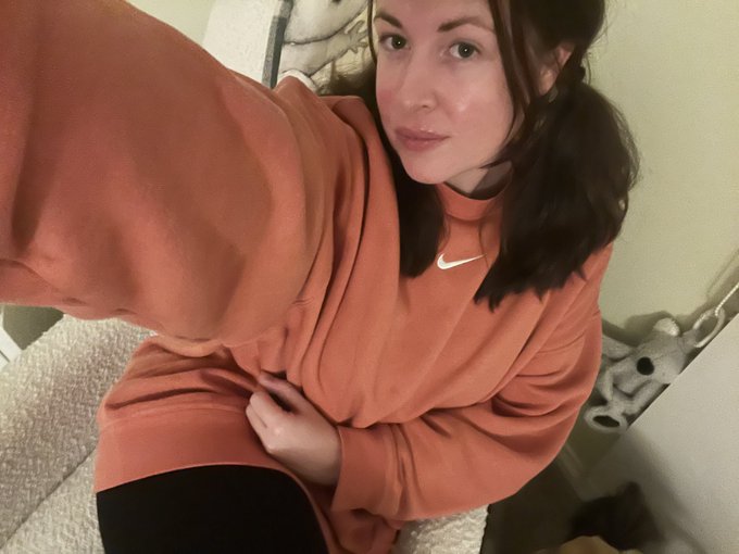 A selfie of a woman wearing an orange Nike sweatshirt in what appears to be a bathroom.