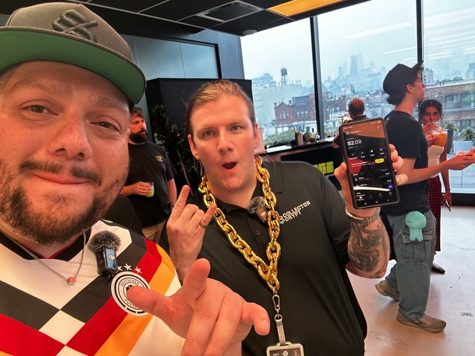 A man wearing a "Collector Crypto" shirt and a thick gold chain holds up his phone displaying the Solflare app, while another man wearing a "$OL" cap poses beside him, captured at the Solana Skyline event in NYC with the city skyline visible through the windows behind them.