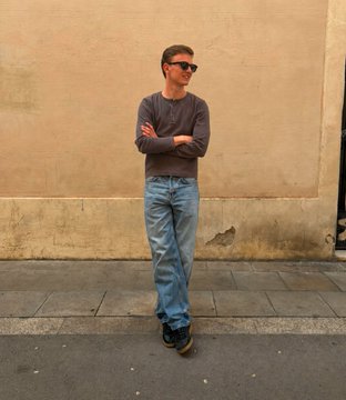 A person standing against a beige wall wearing a gray long-sleeve shirt, blue jeans, and sunglasses, with arms crossed in a relaxed pose.