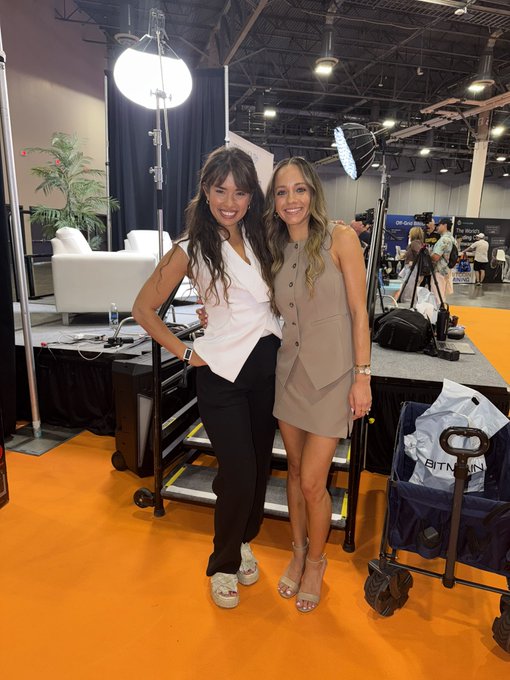 Two women pose together at what appears to be a convention or trade show floor, with lighting equipment and a booth visible behind them.