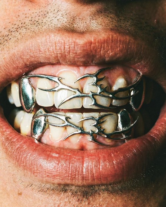 Extreme close-up of a person's mouth displaying ornate silver grillz with intricate branch-like designs covering their teeth.