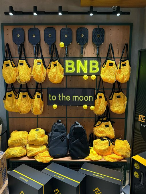 A retail display features BNB-branded pickleball paddles, yellow mesh sports bags, and backpacks arranged on a pegboard wall with illuminated signage reading "BNB to the moon," promoting a Binance-themed pickleball event tied to the post's mention of a Binance Olympic Ball activity that combines crypto trading culture with morning exercise.