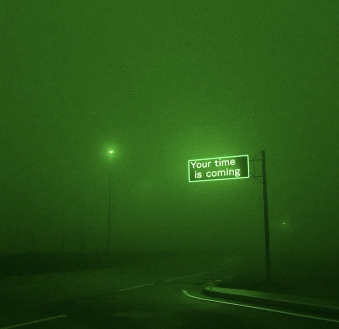 An atmospheric, green-tinted night photograph of an empty road with a glowing neon sign reading 'Your time is coming' and a streetlight in the foggy distance.