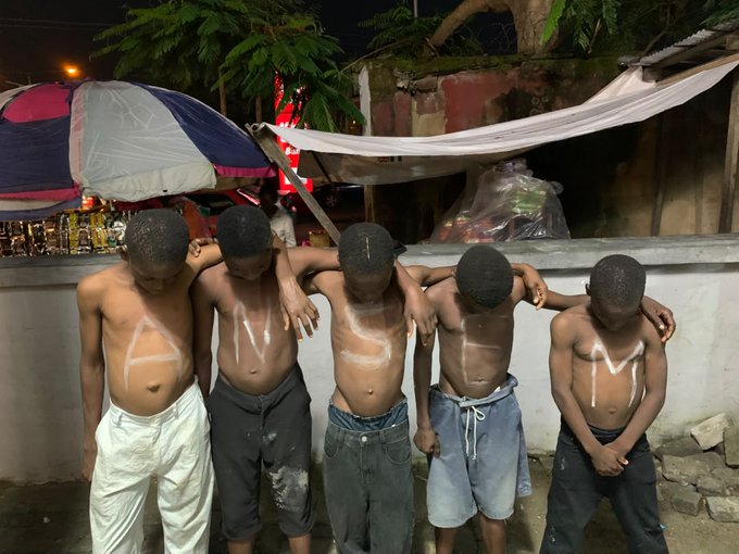 Five shirtless young boys stand with heads bowed, linked arm-in-arm, with white paint spelling "ANSEM" across their torsos against an outdoor nighttime backdrop. The image pairs with the post's caption to show a fan's costly tribute to the crypto influencer $ANSEM, highlighting the lengths supporters will go to in devotion to the meme coin.