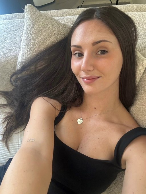 A young woman with long brown hair wearing a black tank top and a heart pendant necklace takes a selfie while lying on a textured cream-colored couch, with a small '25' tattoo visible on her arm.