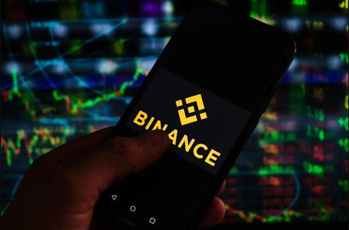 A hand holds a smartphone displaying the Binance cryptocurrency exchange logo, with a background of colorful stock market trading charts.