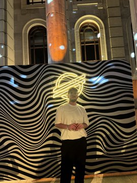 In this image, a man dressed in a white shirt stands against an urban backdrop with an intriguing neon sign featuring a spiral design that seems to be floating above him. The neon sign is illuminated by light and has a logo at its center. The man appears to have his hand on his hip while looking straight ahead towards the camera.

In the background, there are other people in the room attending what appears to be an event or party, with one individual close to the left edge of the image and another standing near the right side. Additionally, a banner is visible at the far right edge of the photo. The man's position seems to be in front of this banner.