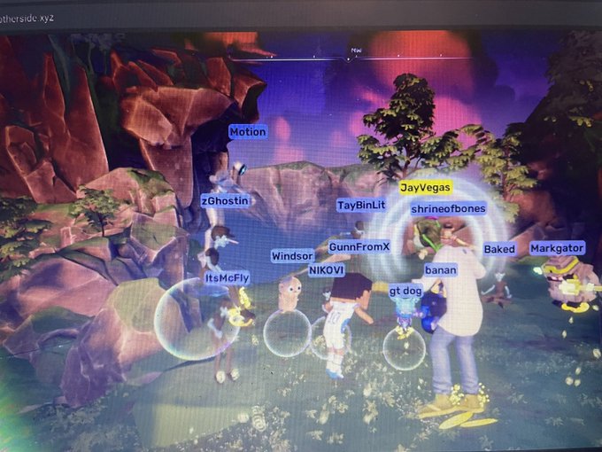 A photo of a computer screen displaying a virtual 3D environment (Metaverse) with multiple avatar players labeled with usernames like 'Motion', 'zGhostin', 'TayBinLit', 'NIKOVi', 'JayVegas', and 'Baked' gathered in a colorful landscape.