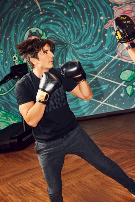 A man in a black t-shirt and boxing gloves trains in a gym with a colorful mural wall, while a partner's glove appears in the frame.