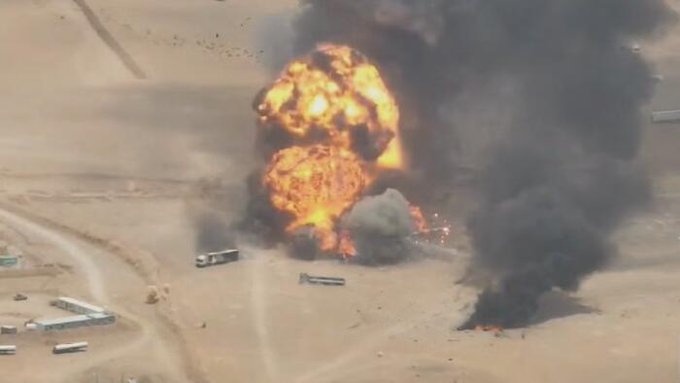 An aerial photograph of a massive explosion in a desert area, with large fireballs and thick plumes of black smoke rising near vehicles and structures on the sandy ground.