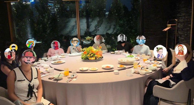 A group of people seated around a large round dining table with various dishes, with the faces of each person covered by different anime and cartoon character stickers.