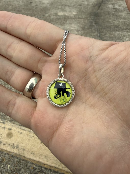 A close-up shows a hand holding a silver pendant on a chain, featuring a yellow circular design with a small character wearing a black hat.